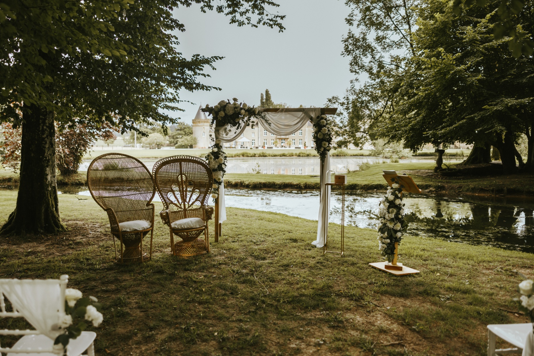 Cérémonie laïque au bord de l'étang du Domaine Château du Faucon, avec arche fleurie et fauteuils en rotin